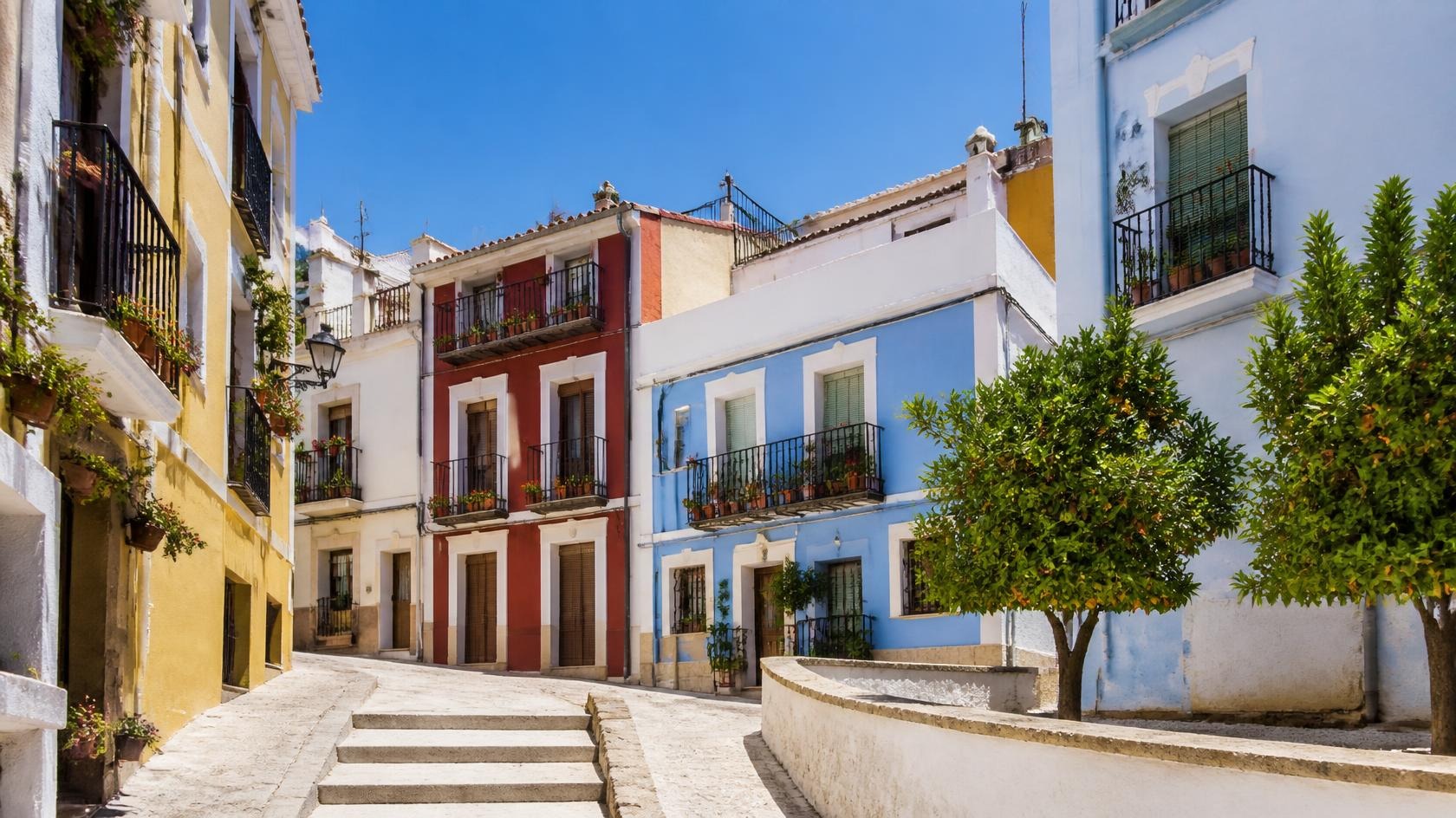 Quiet sunny old-town street in Alicante with colourful houses, balconies with flower pots, a small plaza with orange trees and a clear blue sky