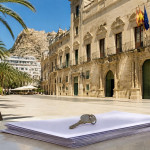 House key and a stack of papers on a café table in a sunny Alicante square, with palm trees and the castle behind
