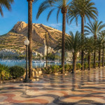 Sunny autumn morning on Alicante's palm-lined Explanada promenade with its wavy mosaic tiles, the marina and the Mediterranean, and Santa Bárbara Castle on Mount Benacantil under a blue sky