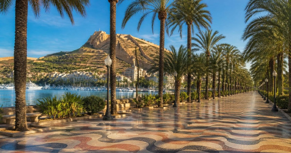 Sunny autumn morning on Alicante's palm-lined Explanada promenade with its wavy mosaic tiles, the marina and the Mediterranean, and Santa Bárbara Castle on Mount Benacantil under a blue sky